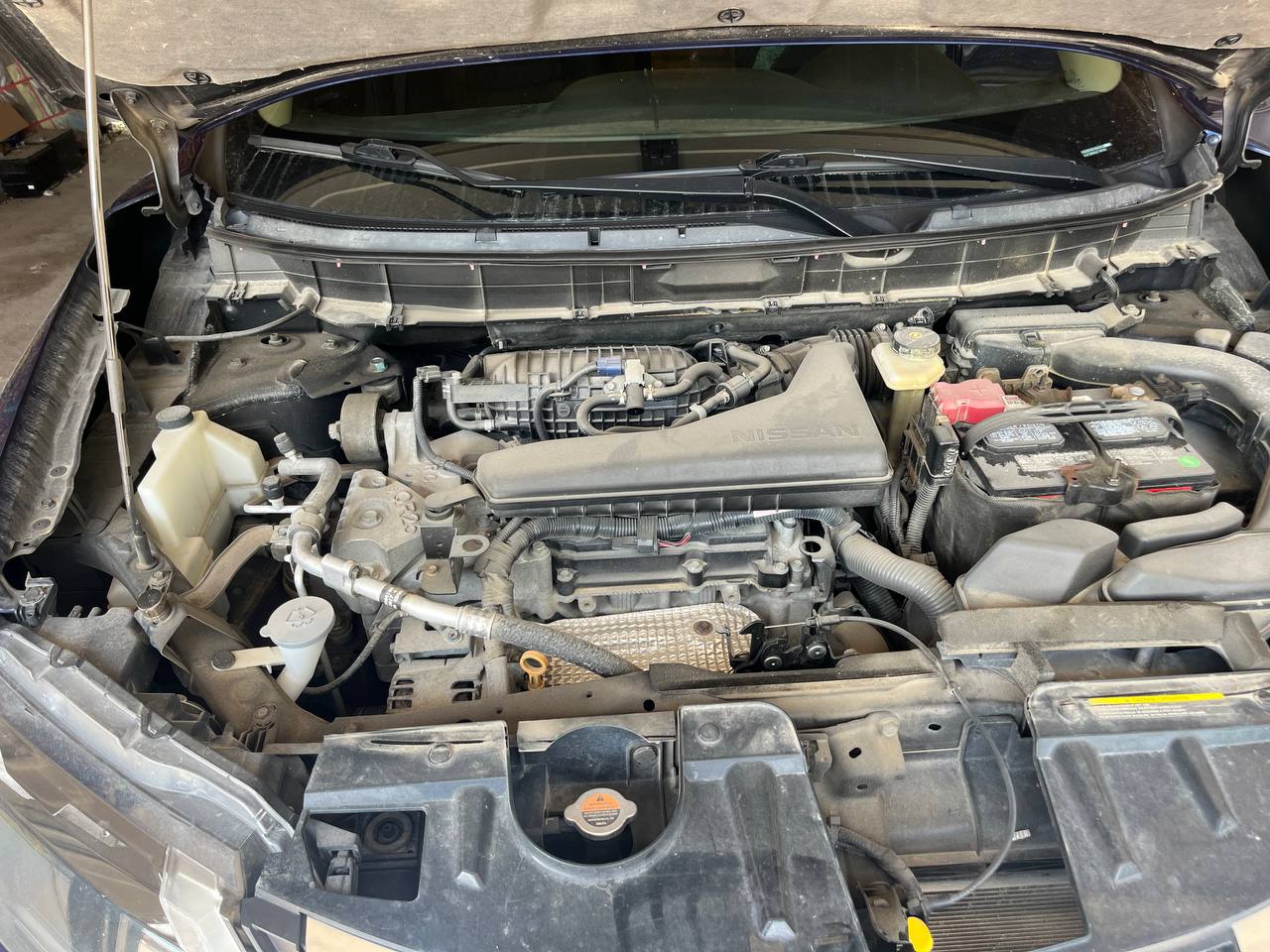 Engine bay of a blue Nissan Rogue before cleaning, with dust and grime settled across the intake cover, battery and surrounding plastics