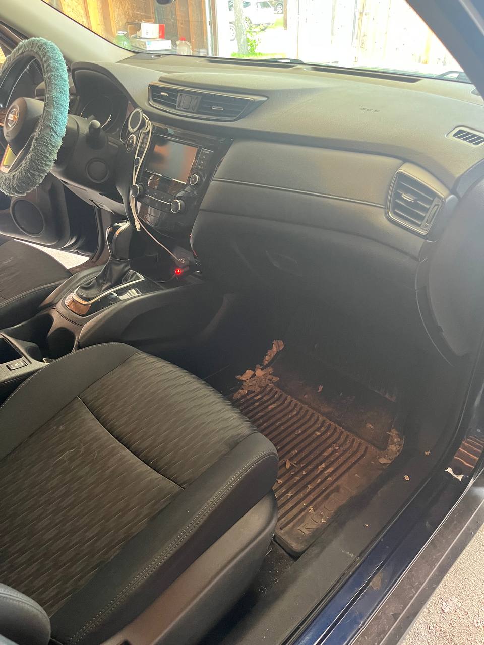 Passenger footwell of a blue Nissan Rogue before cleaning, with dead leaves and dried mud across the rubber mat and a dusty dash above it