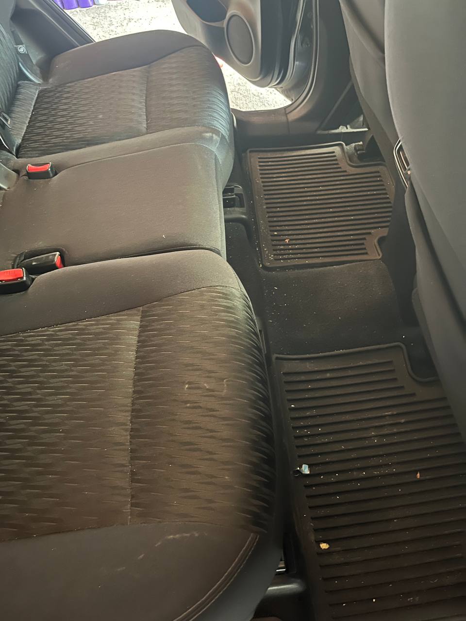 Rear bench and rear footwells of a blue Nissan Rogue before cleaning, with crumbs and debris across the cloth seats and mats