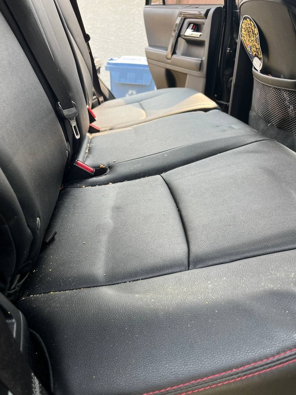 Rear bench of the same 4Runner before cleaning, with crumbs across the black leather and collected along the seams and seatbelt buckles